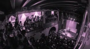 A crowded concert venue with balconies and a stage. People are watching a performance under purple lighting.